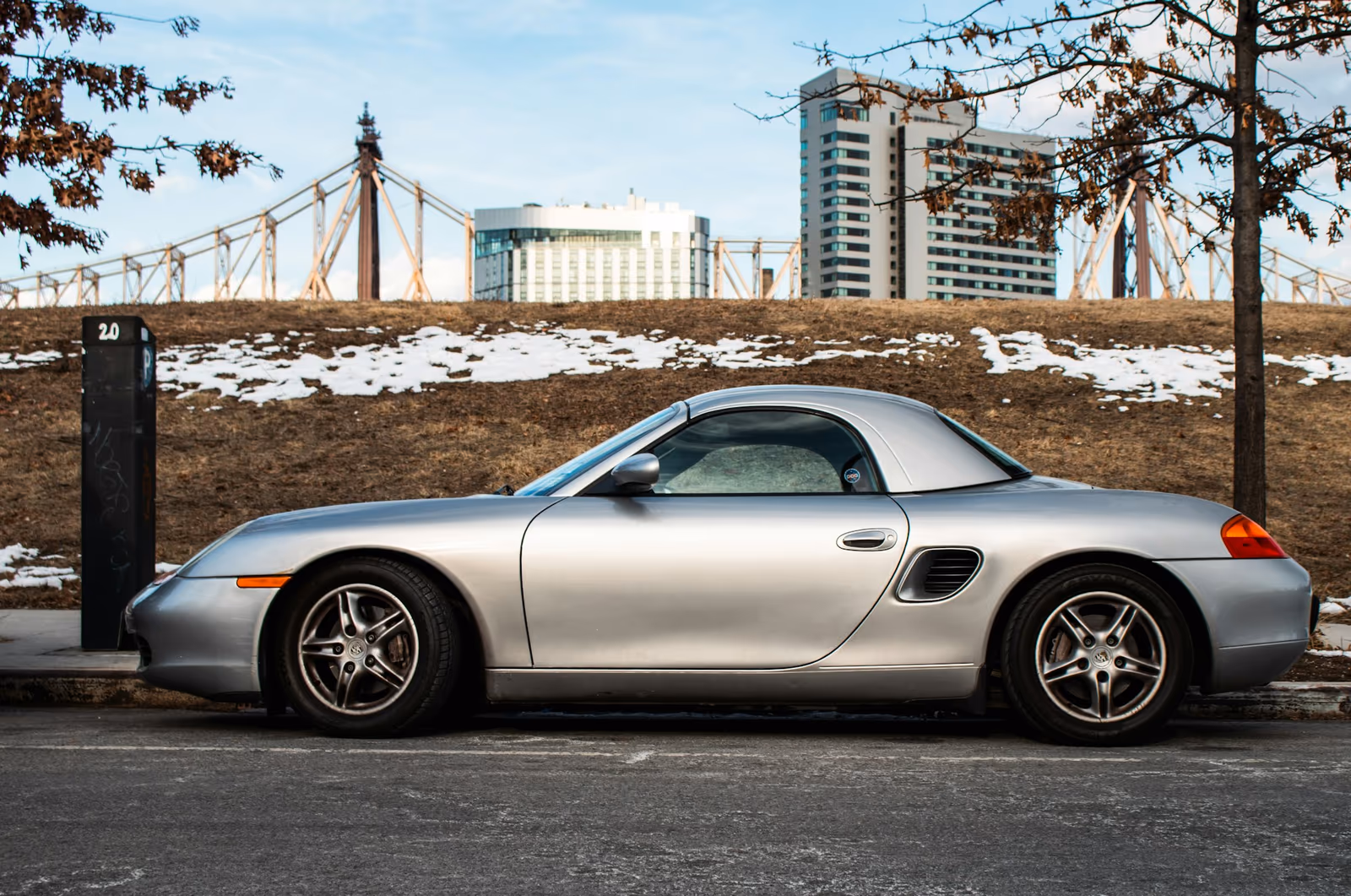 Silberner Porsche Boxster 986 mit Hardtop am Straßenrand vor einer Brücke, Seitenansicht